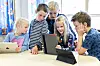 Barneskoleelever står og sitter rundt et bord og bruker nettbrett i et klasserom.