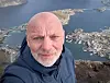 Mann i mørk jakke tar selfie med havn og øyer bak seg.