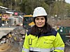 Person i gul refleksjakke og hvit hjelm foran en anleggsplass med gravemasser og maskiner.