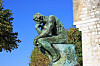 Bronseskulptur av en tenkende figur på sokkel ved en steinmur under blå himmel.
