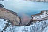 Snødekt elvebredd ved et stille vann med bratte fjellsider og is langs kanten.