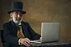 Eldre mann i hatt sitter ved et bord og jobber på en laptop mot mørk bakgrunn.