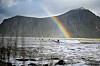 Surfer på en bølge under en regnbue med fjell og grått vann i bakgrunnen.