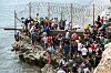Mange mennesker står tett ved et piggtrådgjerde på en steinete kyst mens de prøver å ta seg mot Ceuta.