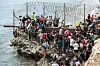 Mange mennesker står tett ved et piggtrådgjerde på en steinete kyst mens de prøver å ta seg mot Ceuta.