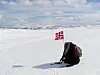 Norsk flagg i snøen ved en ryggsekk med fjell under blå himmel.