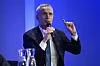 Jens Stoltenberg taler i mikrofon mot en blå bakgrunn, med glass på bordet foran seg.