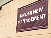 Lilla skilt med teksten «UNDER NEW MANAGEMENT» på en lys butikkvegg.
