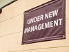 Lilla skilt med teksten «UNDER NEW MANAGEMENT» på en lys butikkvegg.