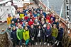 Stor gruppe mennesker poserer på skipsdekket med sjøen i bakgrunnen.