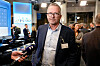 Geir Røsvoll i dress står foran pressen i en møtesal med skjermer og mennesker i bakgrunnen.