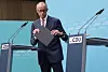 Friedrich Merz står mellom CDU-talerstoler og holder en mørk mappe foran en blå bakgrunn.