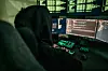 Hettekledd person arbeider ved flere dataskjermer med grønn kode.