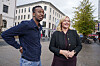 To personer står sammen på et byfortau, med bygninger, trær og trafikk i bakgrunnen.