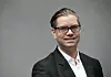 Portrett av Marius Gonsholt Hov i mørk dress og briller mot en grå bakgrunn.